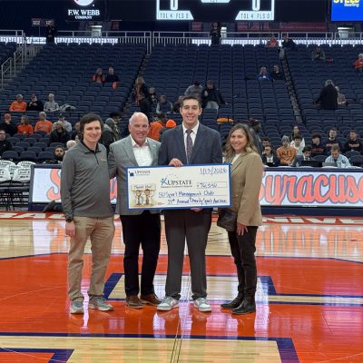 Four people standing on a basketball court holding an oversized donation check during a Syracuse University game, with stadium seating and spectators in the background