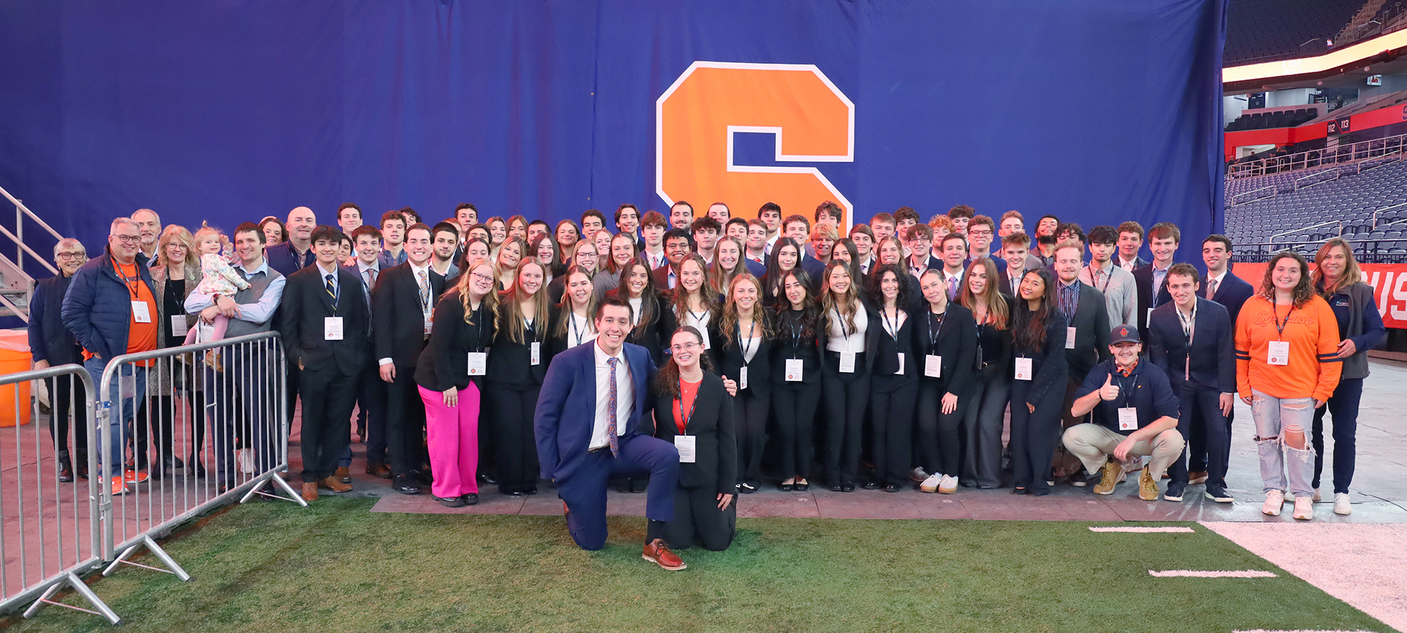 A large group of students and faculty posing together on a stadium field in front of a blue backdrop with an orange ‘S’ logo