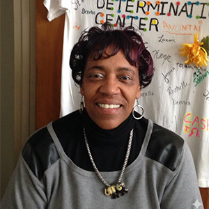 A woman in a black and grey shirt with a gold necklace, smiling. A sign that says Determination Center is behind her.