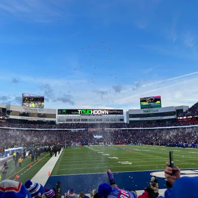 Wide-angle view inside a packed football stadium during a Buffalo Bills game, with the end zone and goalposts in the foreground, fans in blue and red filling the stands, confetti floating in the air, and large video boards displaying “Touchdown” under a bright blue winter sky.