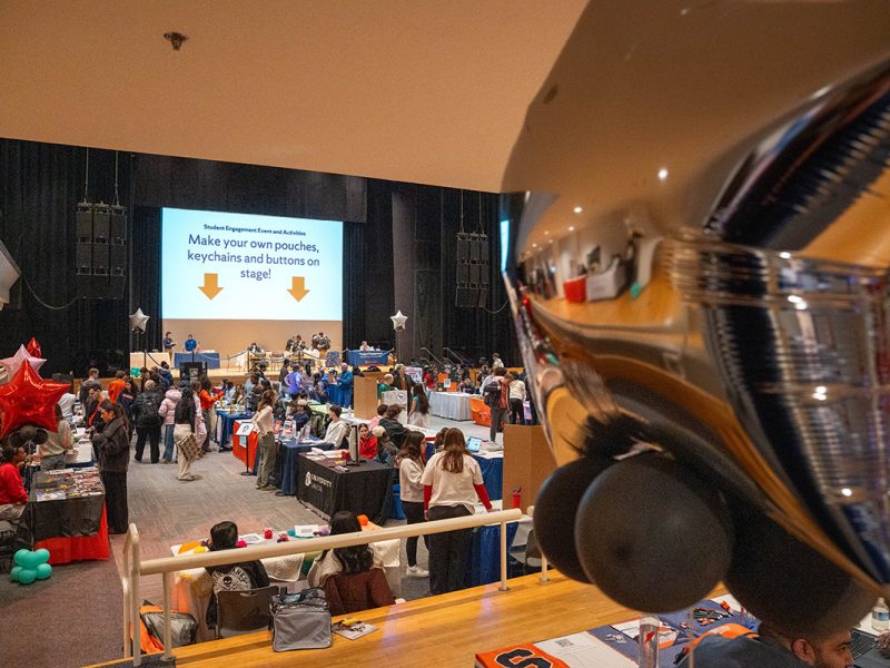 Large indoor involvement fair with rows of tables, colorful balloons, and attendees exploring booths, with a stage in the background displaying a sign about making custom pouches, keychains, and buttons.