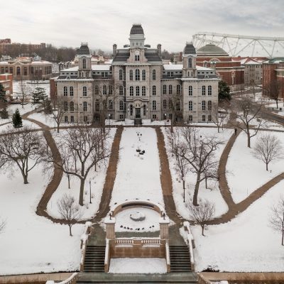 A large historic building with four towers stands at the center of a snow-covered campus, surrounded by leafless trees and curved walkways.