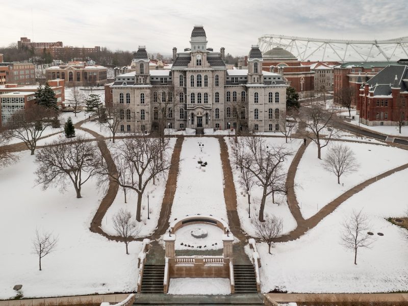 A large historic building with four towers stands at the center of a snow-covered campus, surrounded by leafless trees and curved walkways.