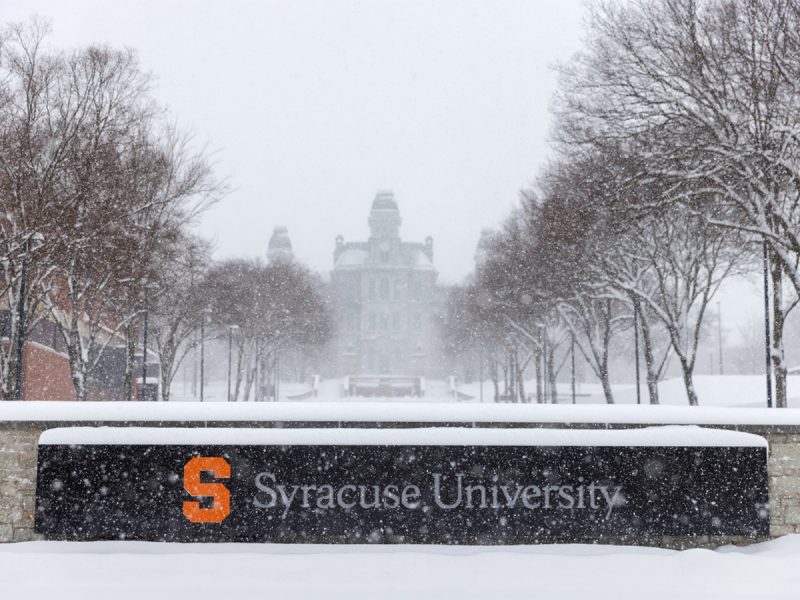 Snow-covered Syracuse University entrance sign with trees lining the walkway and the Hall of Languages visible in the background during a winter snowfall
