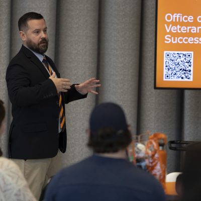 Presenter speaks to a small seated audience beside a screen displaying “Office of Veteran Success” with a QR code.