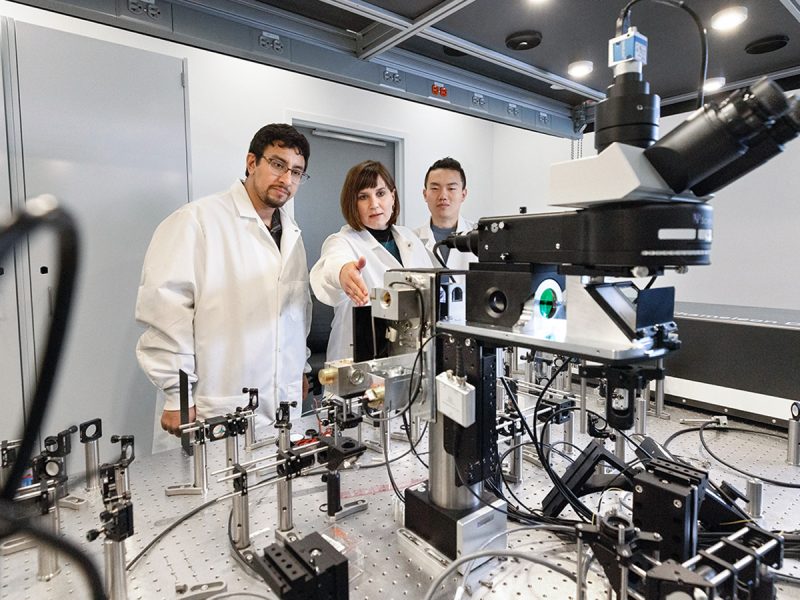Three people--two students and a professor--in a lab, wearing white lab coats. They are pictured with lab equipment.