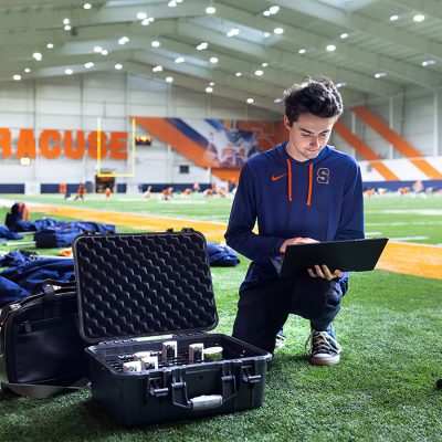 Student kneeling on turf at the Ensley Center. Working on a laptop with equipment nearby