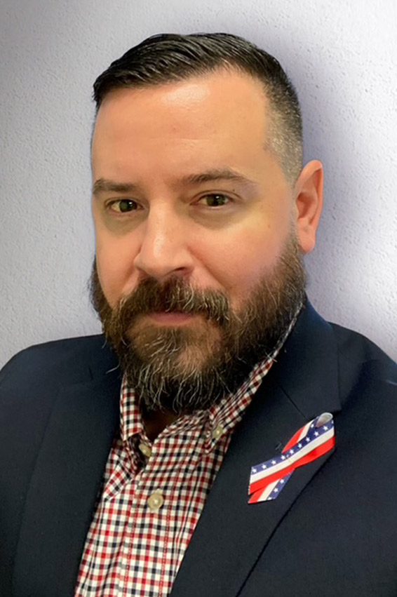 Head-and-shoulders portrait of a person in a blazer and checkered shirt with a small patriotic ribbon pin on the lapel.