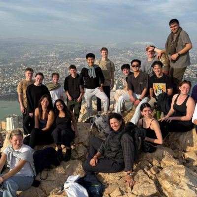 students pose on rocky overlook above coastal city with mountains and bay in background.