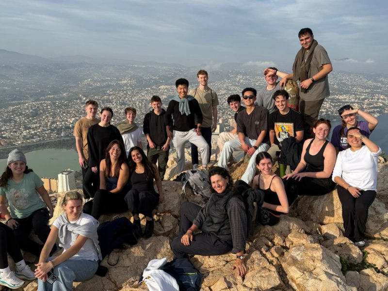 students pose on rocky overlook above coastal city with mountains and bay in background.