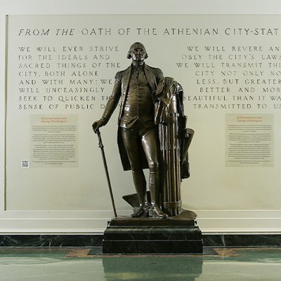 Bronze statue of George Washington standing with a cane beside a column, displayed against a wall featuring text from the Athenian Oath.
