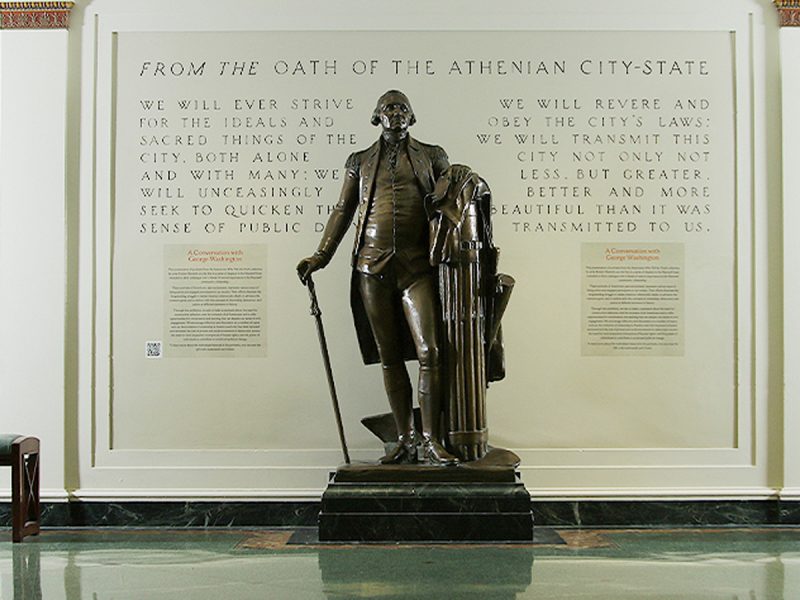 Bronze statue of George Washington standing with a cane beside a column, displayed against a wall featuring text from the Athenian Oath.