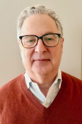 Person with gray curly hair wearing a light-colored collared shirt and a rust-colored V‑neck sweater.