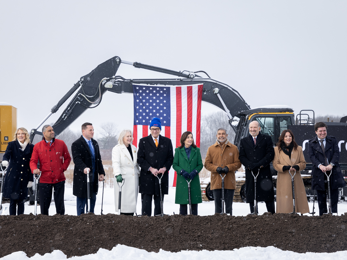 Officials and dignitaries holding ceremonial shovels stand in front of construction equipment and a large American flag during the Micron Technology groundbreaking ceremony in snowy conditions.