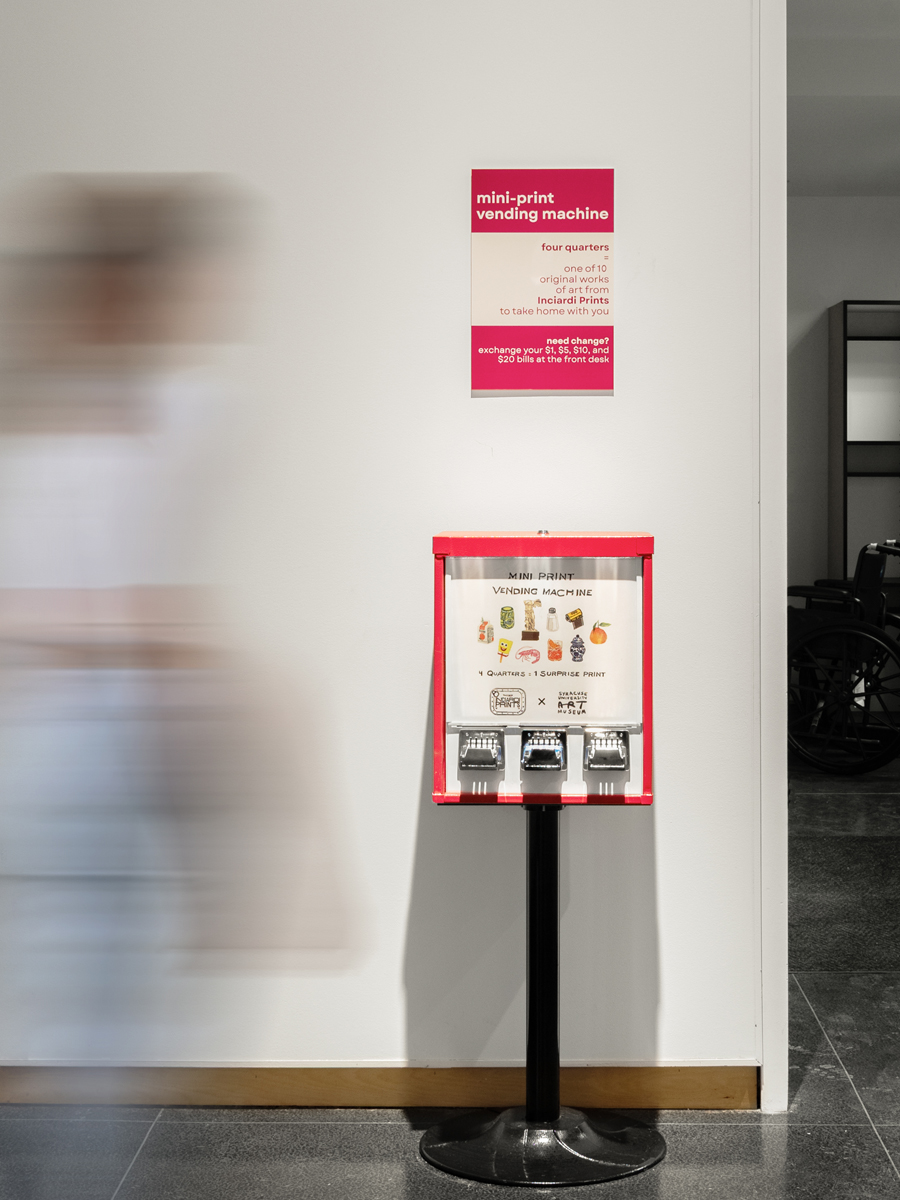 A bright red mini-print vending machine stands on a black pedestal in a white-walled gallery space.