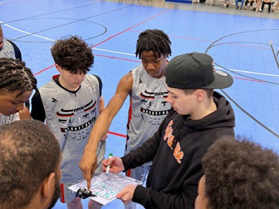 Basketball players in grey and black jerseys strategize with a coach during a game.
