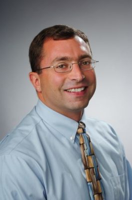 A person smiles while posing for a headshot in front of a grey backdrop.