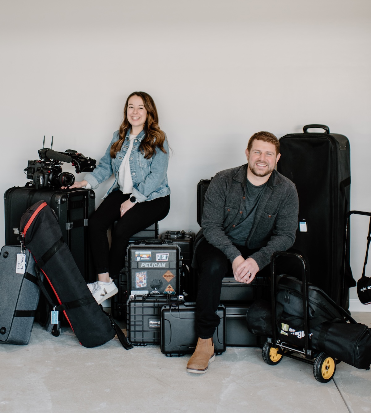 Two people sit among an assortment of professional camera gear, equipment cases, and travel bags in a minimalist room.
