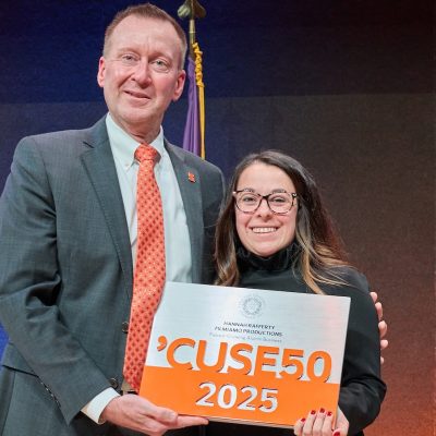 Two people standing on a stage hold a sign that reads “’CUSE50 2025,” with university flags displayed behind them.