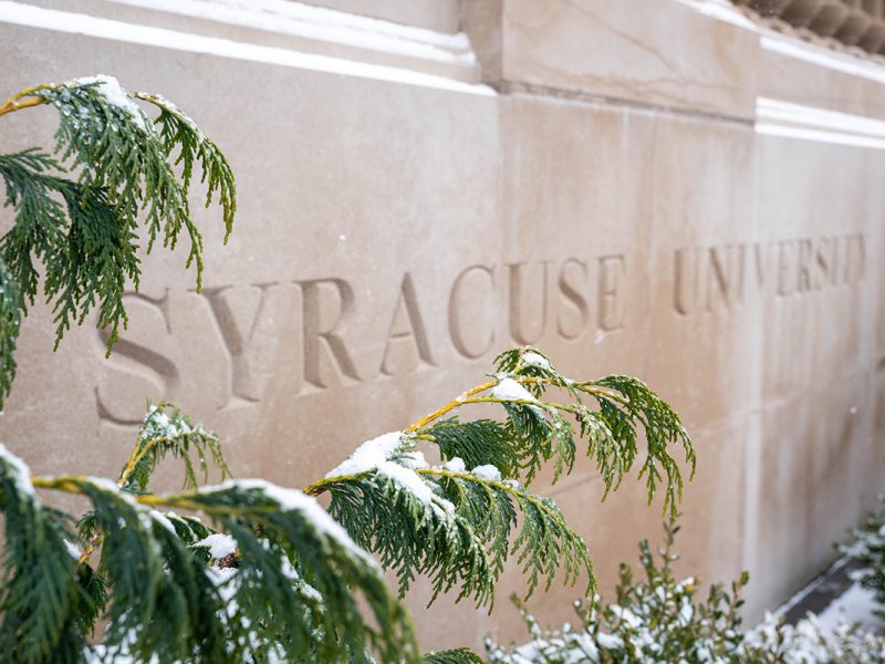 Snow-covered evergreen branches in front of a stone wall engraved with ‘Syracuse University.’