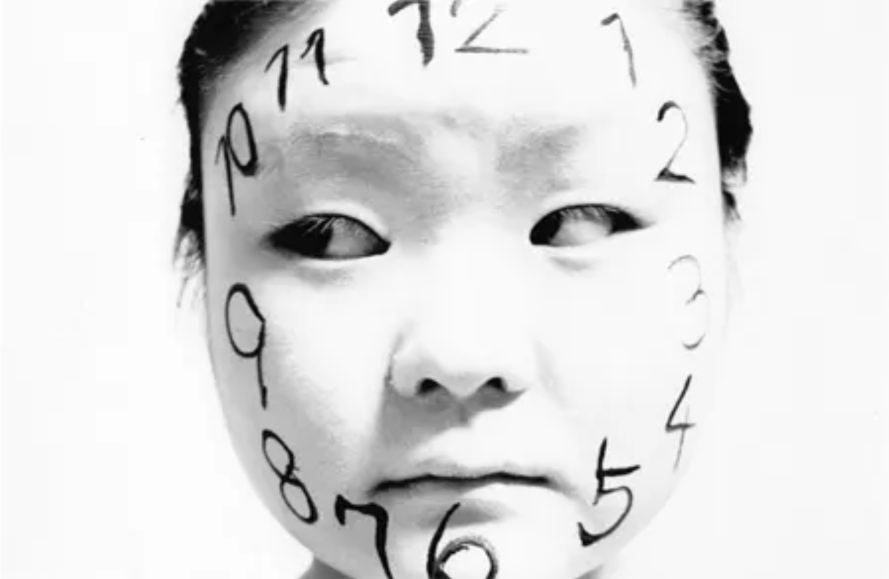 Black and white self-portrait photograph showing a face with clock numerals painted across it.