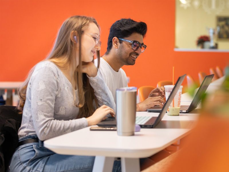 Two people working on laptops at a modern table with orange chairs and a bright orange wall in the background; stainless steel tumbler and iced drink on the table.