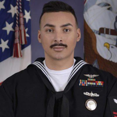 U.S. Navy service member in dress uniform seated in front of American and Navy flags