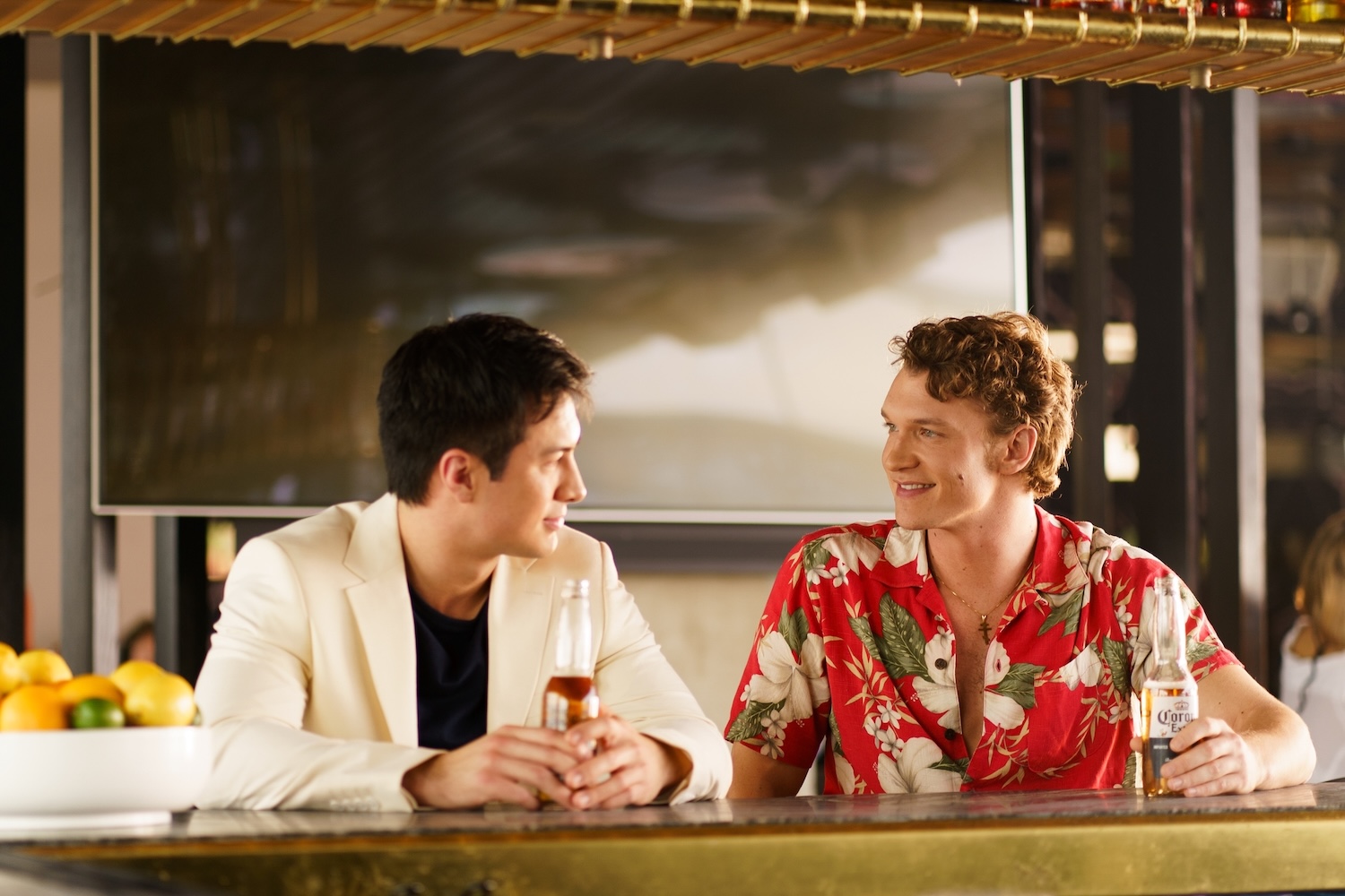 Two people sit at a bar, each holding a beer, talking in a relaxed, tropical-themed setting.
