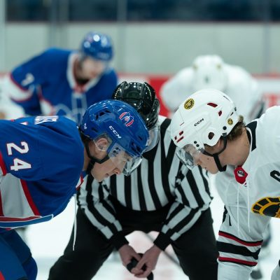 Two hockey players from opposing teams crouch for a faceoff while a referee prepares to drop the puck.