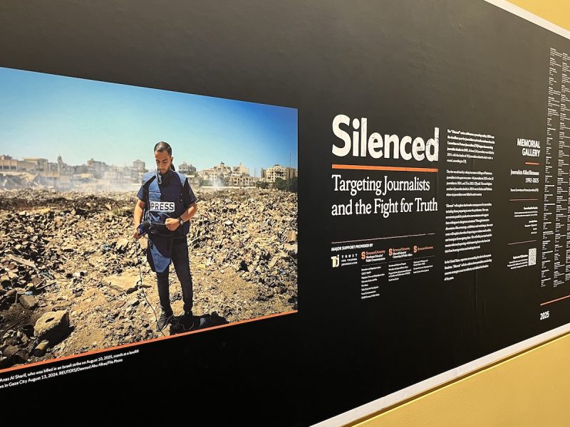 Exhibit panel featuring a large photo of a journalist wearing a press vest in a rocky landscape, alongside text titled “Silenced: Targeting Journalists and the Fight for Truth.”