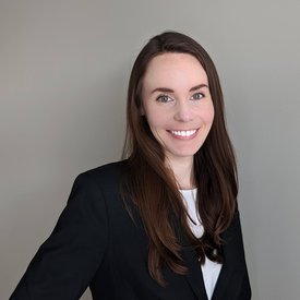 Person with long brown hair wearing a black blazer and white top, standing against a plain gray background