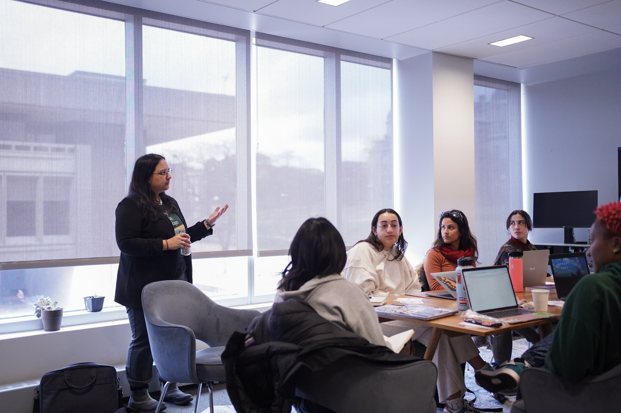 A woman with long dark hair and glasses stands and speaks to a small group of students seated around a table with laptops in a modern, light-filled classroom or studio space with large windows.