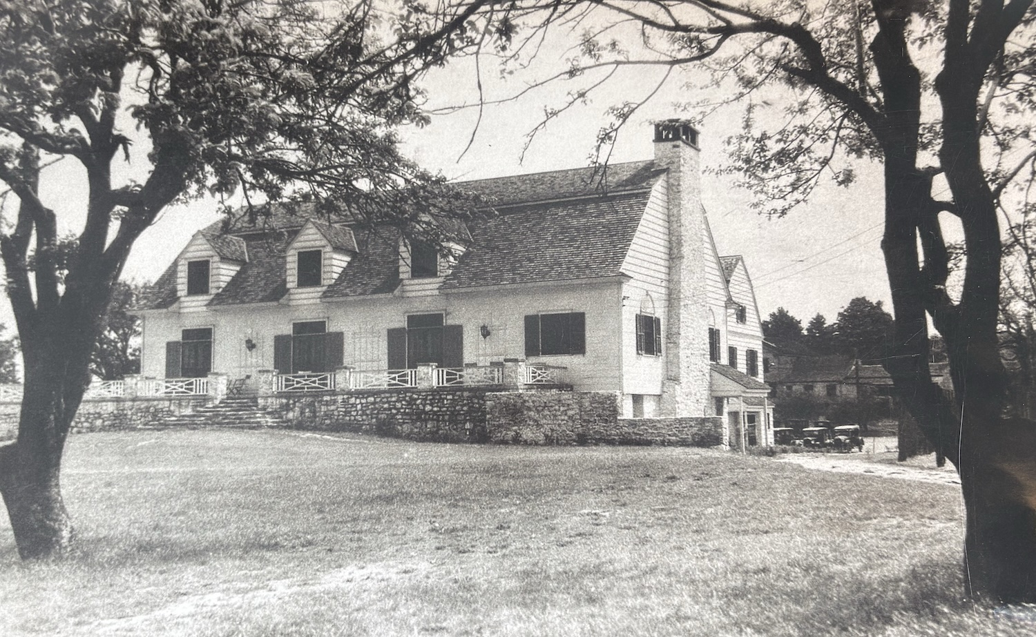 A historic lodge-style building with dormer windows and a stone foundation, framed by large trees.