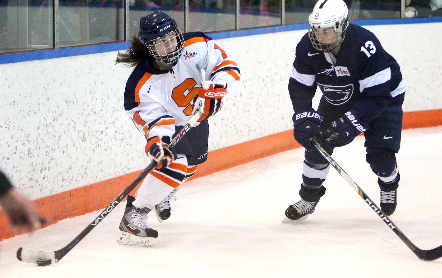 A player in an orange‑and‑white uniform controls the puck while a defender in a dark uniform skates alongside.