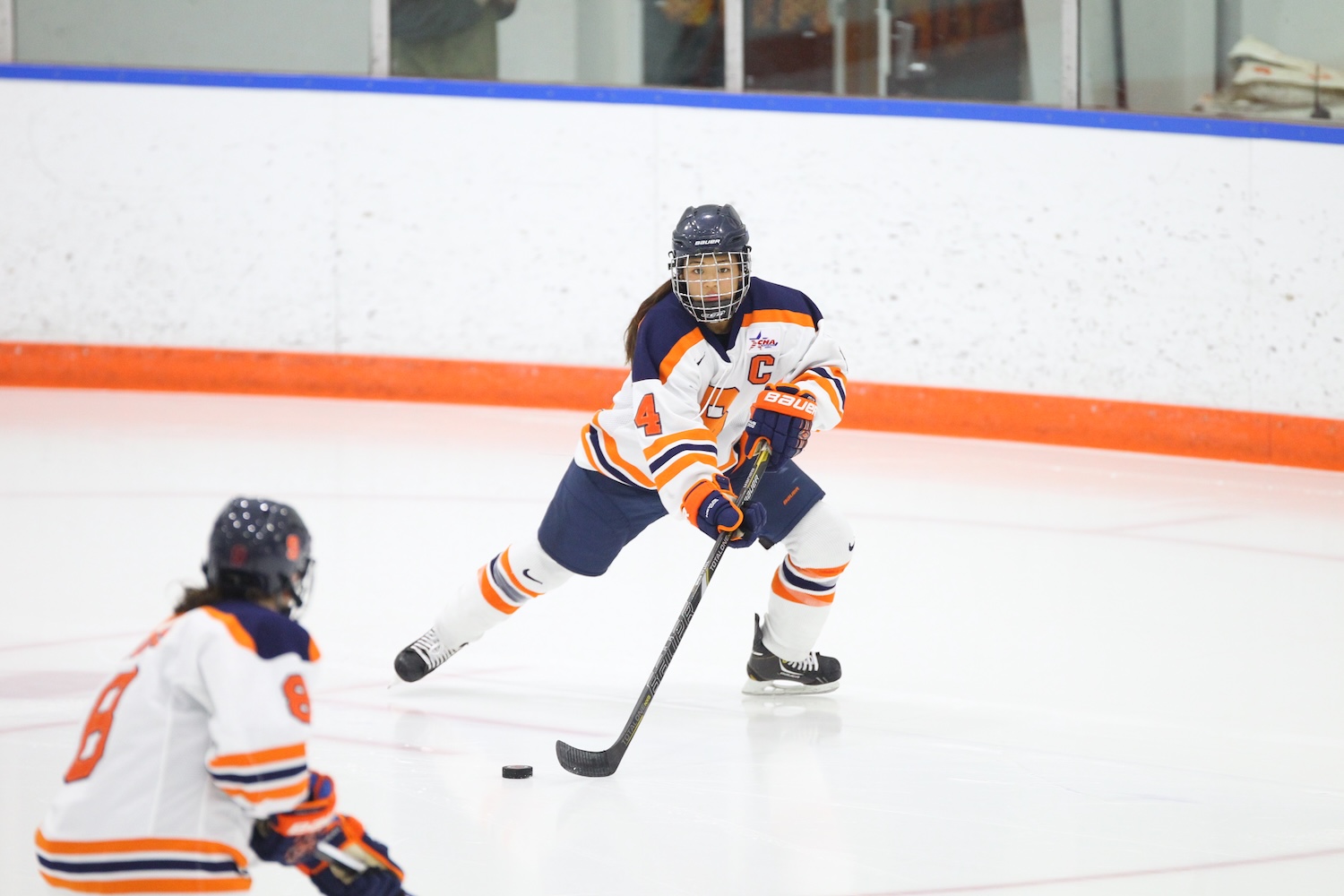 A hockey player in an orange‑and‑white uniform prepares to pass the puck while a teammate skates nearby.