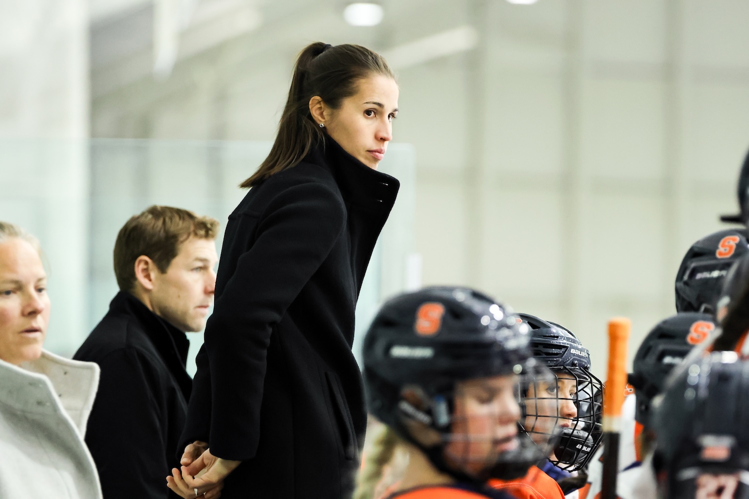 A coach in a black coat stands behind the bench, watching a group of hockey players in dark helmets and orange‑and‑white uniforms. 