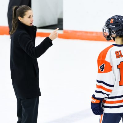 A coach in dark clothing gives instructions to a hockey player wearing an orange‑and‑white uniform with number 14 during a game.