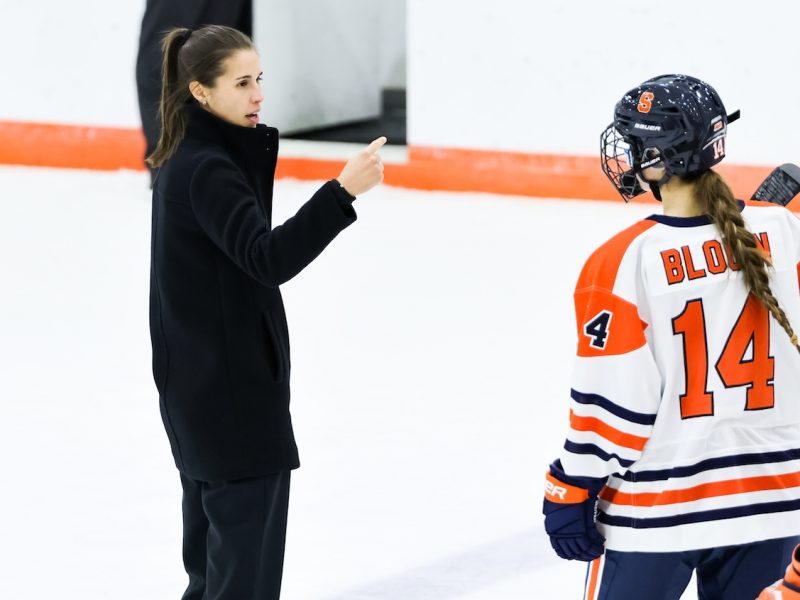 A coach in dark clothing gives instructions to a hockey player wearing an orange‑and‑white uniform with number 14 during a game.