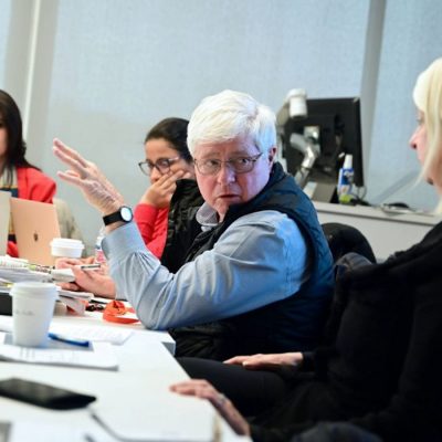 People seated around a conference table engaged in discussion, with laptops, papers, and drinks in front of them.