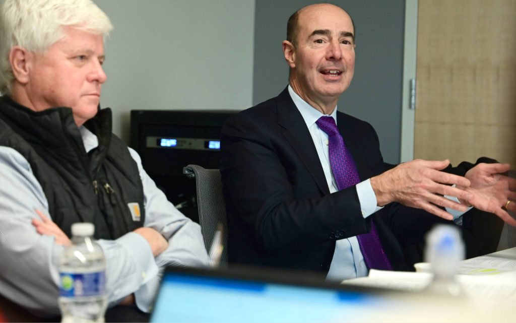 Two individuals in a meeting room talking, with one gesturing while speaking and papers on the table.