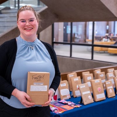 A person holds a book covered in brown paper, standing beside a table with brown paper wrapped-books on display