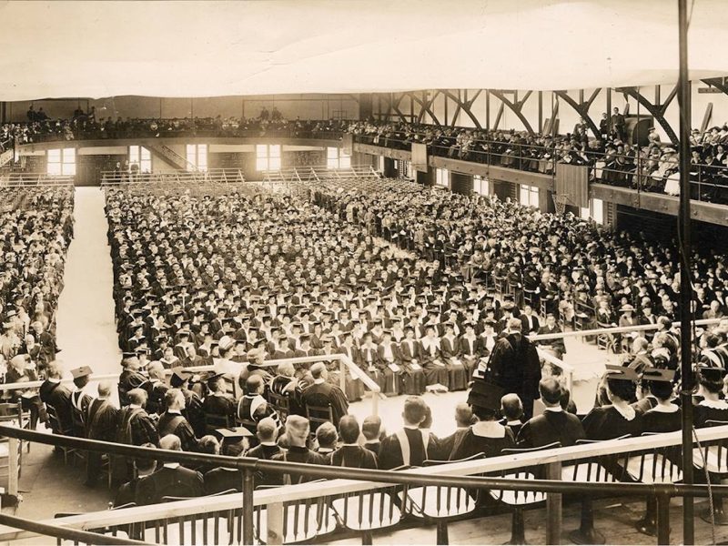 Historic indoor graduation ceremony in a large hall, with hundreds of people seated in rows wearing caps and gowns, surrounded by balconies filled with spectators.
