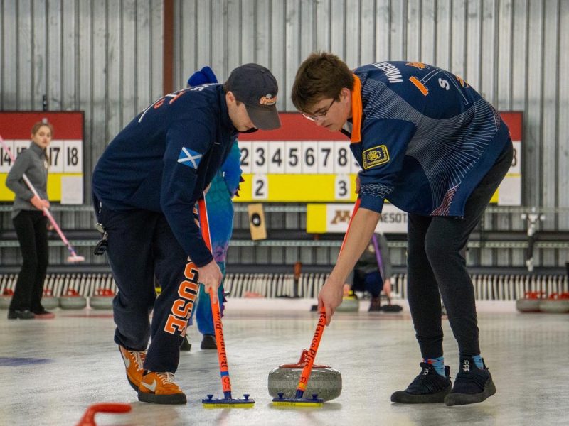 Two curlers in Syracuse University uniforms sweep the ice during competition, guiding a stone toward the target.