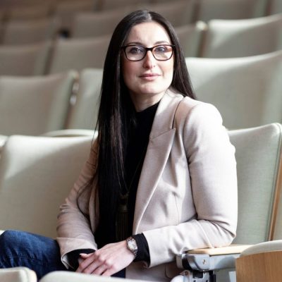A person sits in an empty auditorium-style room, wearing a light blazer.