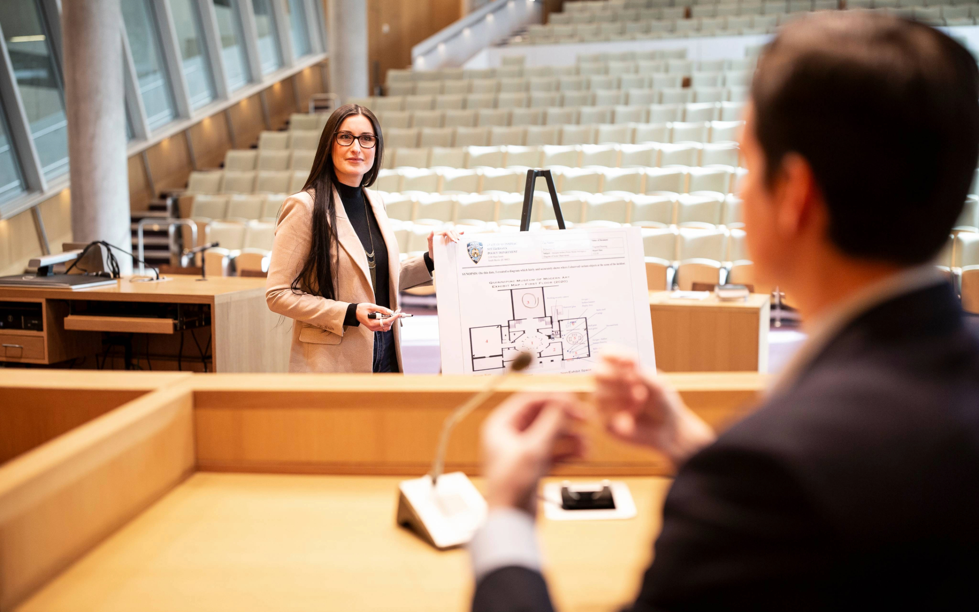 A person stands in a courtroom holding a large diagram on an easel while another person sits at the judge’s bench in the foreground.