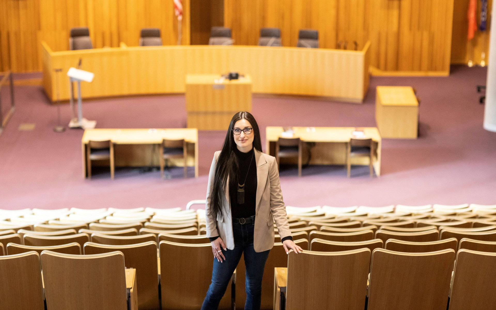 A person stands in the seating area of a large wood-paneled courtroom, facing the camera with the judge’s bench and desks visible in the background.