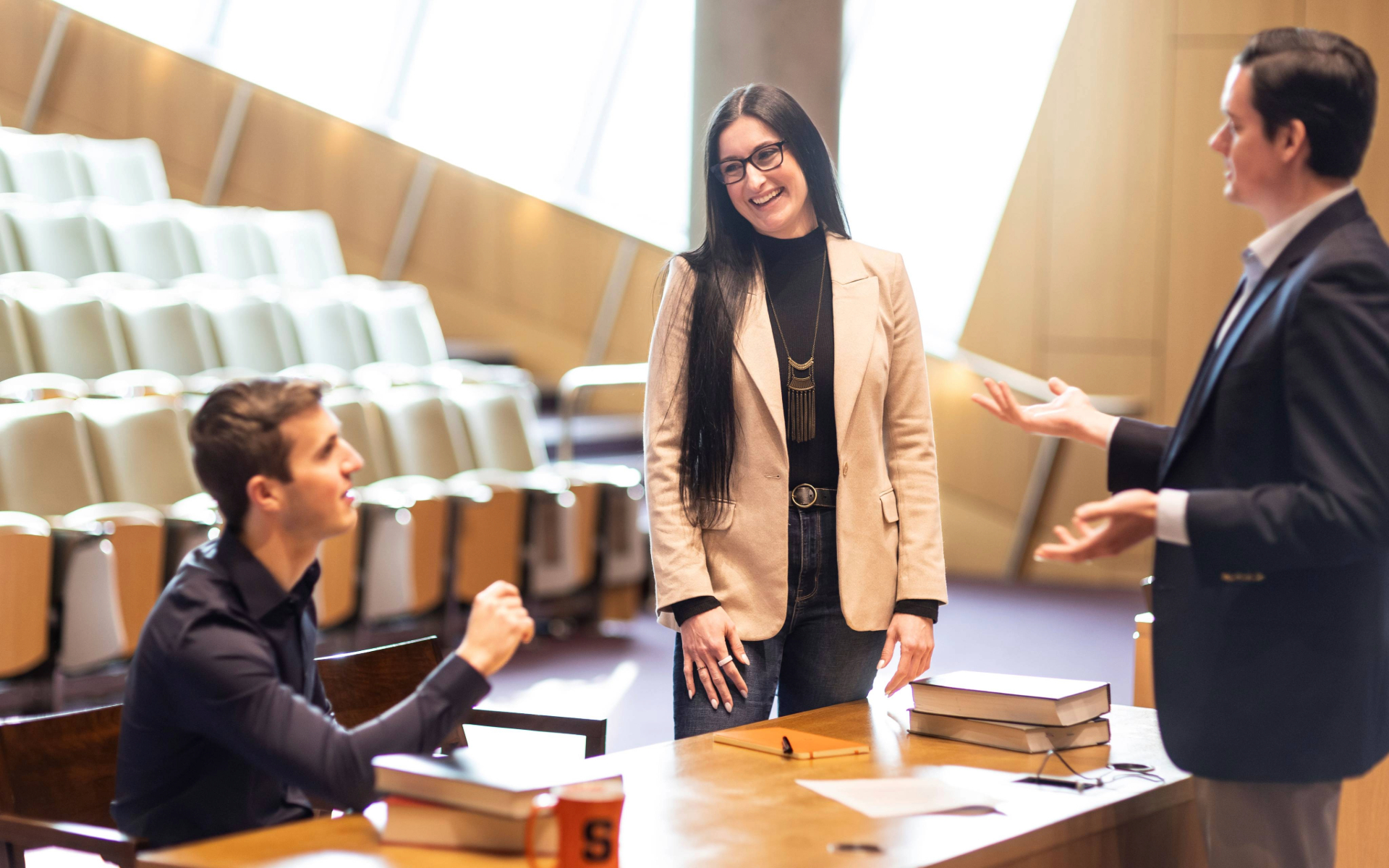 A person stands at a table in a courtroom while two others seated nearby engage in discussion, with books and papers on the table.
