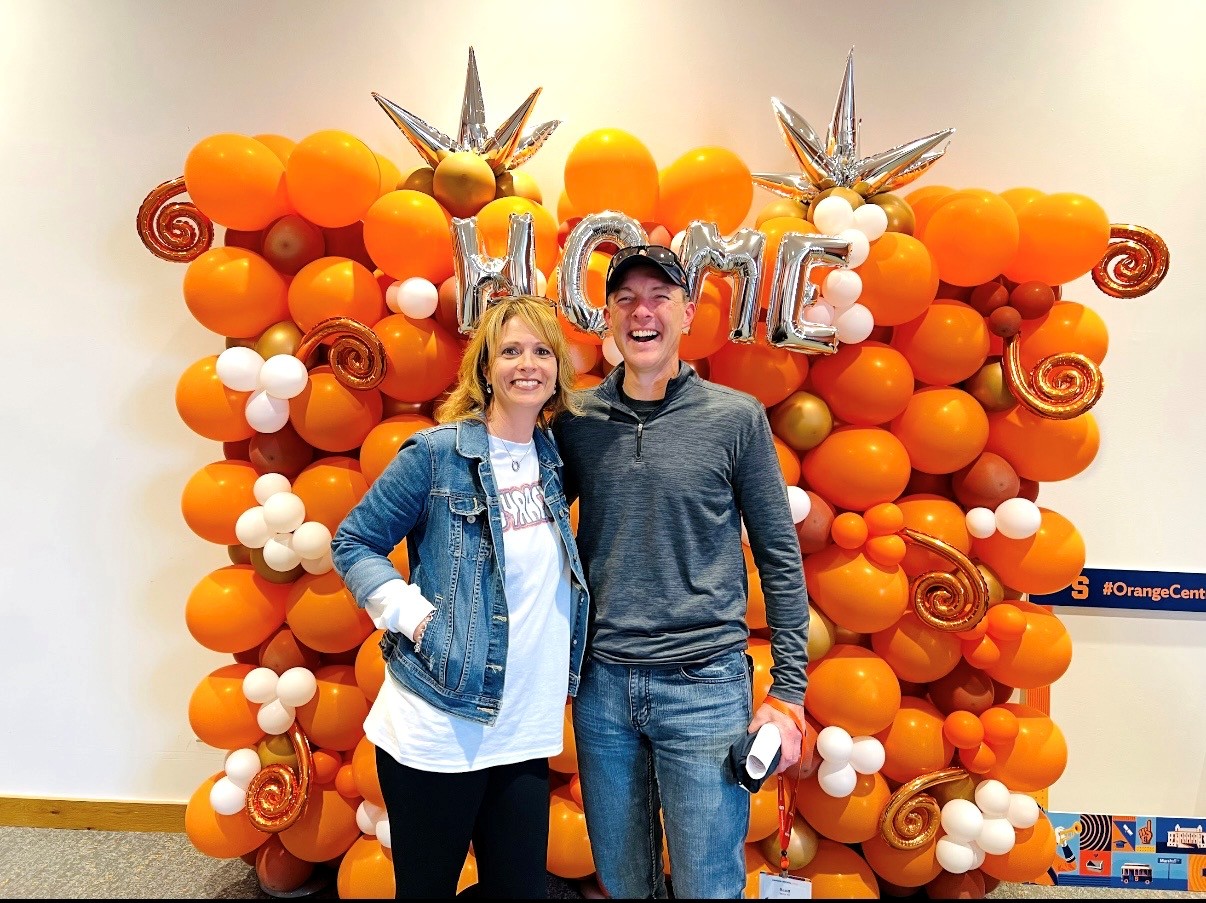 Two people standing in front of a large orange balloon backdrop with silver letters spelling ‘HOME'