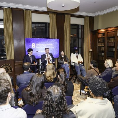 Four panelists speak to an audience during Syracuse University's Center for the Creator Economy event in a wood-paneled room with blue seating and a screen displaying the university logo.
