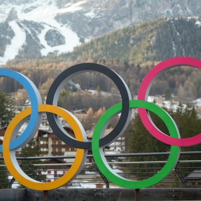 Colorful Olympic rings displayed outdoors with snow‑covered mountains in the background.